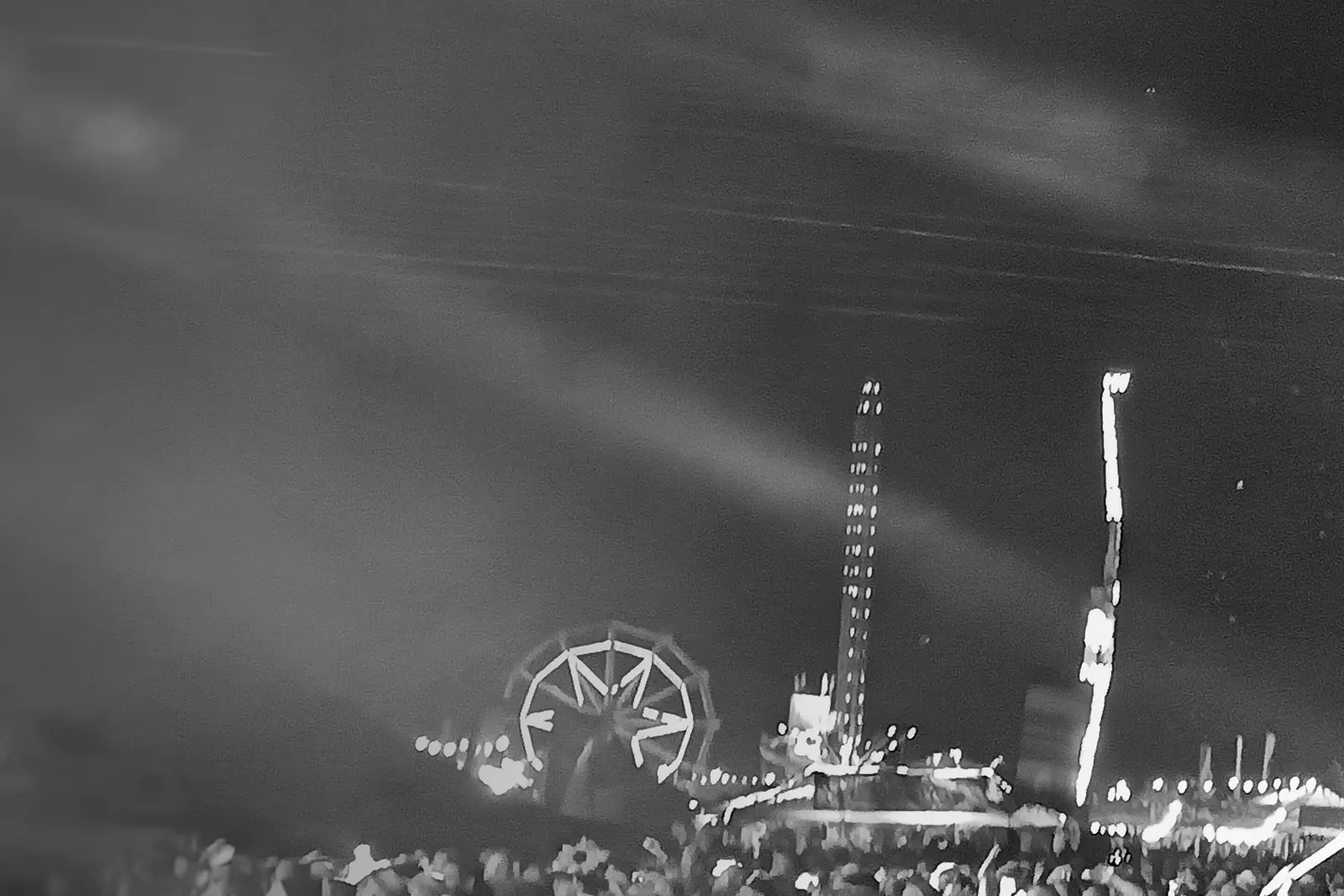 Black and white photo of a festival site at night: light beams across the sky, a lit ferris wheel, a drop tower and rides, with the back of a distant crowd along the bottom edge.