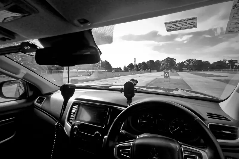 Black and white photo from the driving seat of a site vehicle, looking out through the windscreen across open ground.