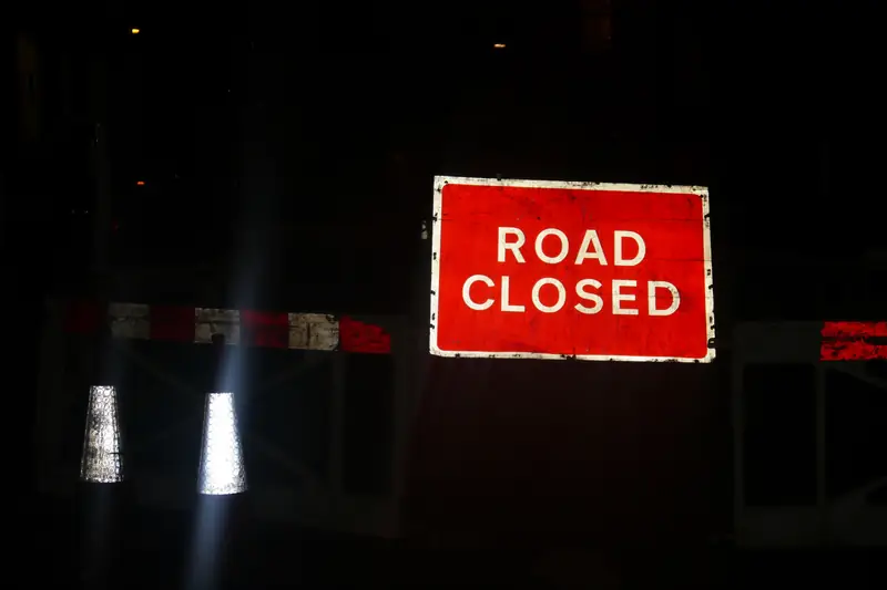 A red Road Closed sign lit up at night across a road, with a barrier and reflective posts below it.