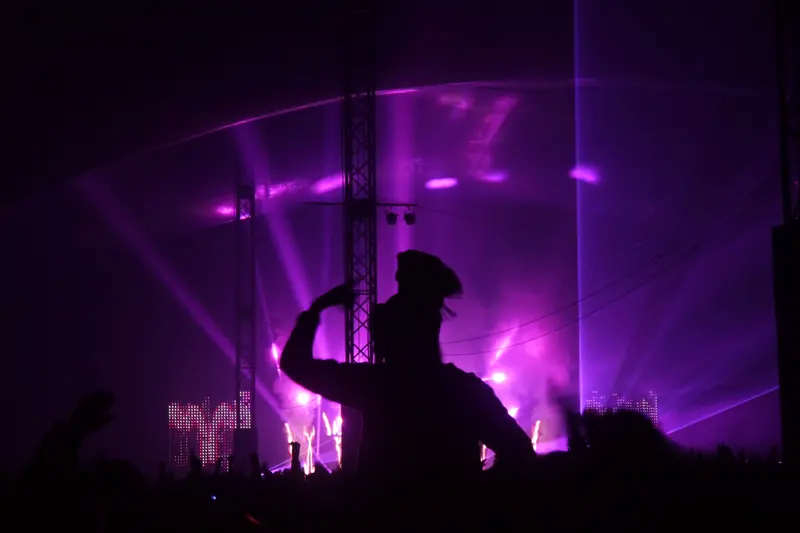 A fan silhouetted against purple stage lights and laser beams in front of a festival stage at night.