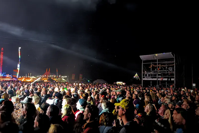 A festival crowd at night watching the main stage, with fairground lights beyond.
