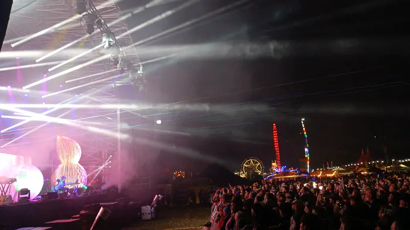 Light beams sweep over a festival crowd at night, with the main stage lit purple on the left and a lit ferris wheel in the distance.