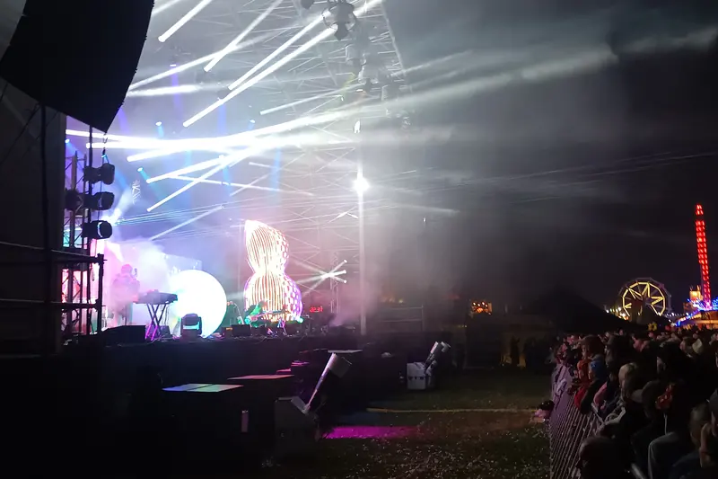 A festival stage lit with beams and haze, with fairground rides lit up beyond the crowd at night.