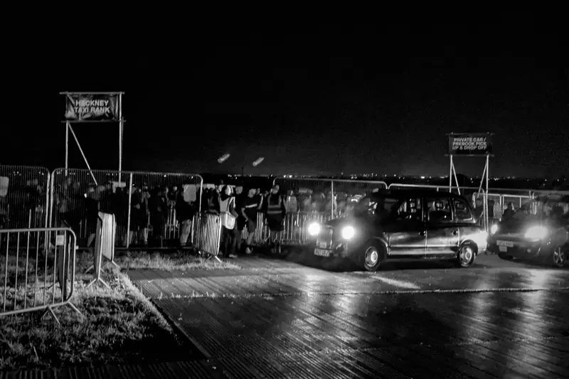 Black and white photo of a site lit by floodlights at night, with vehicles parked beyond the fencing.