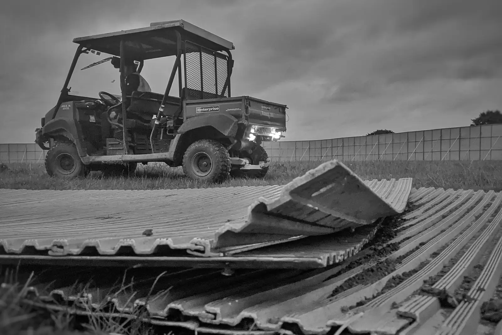 Black and white photo of a utility vehicle on a festival site beside stacked fence panels.